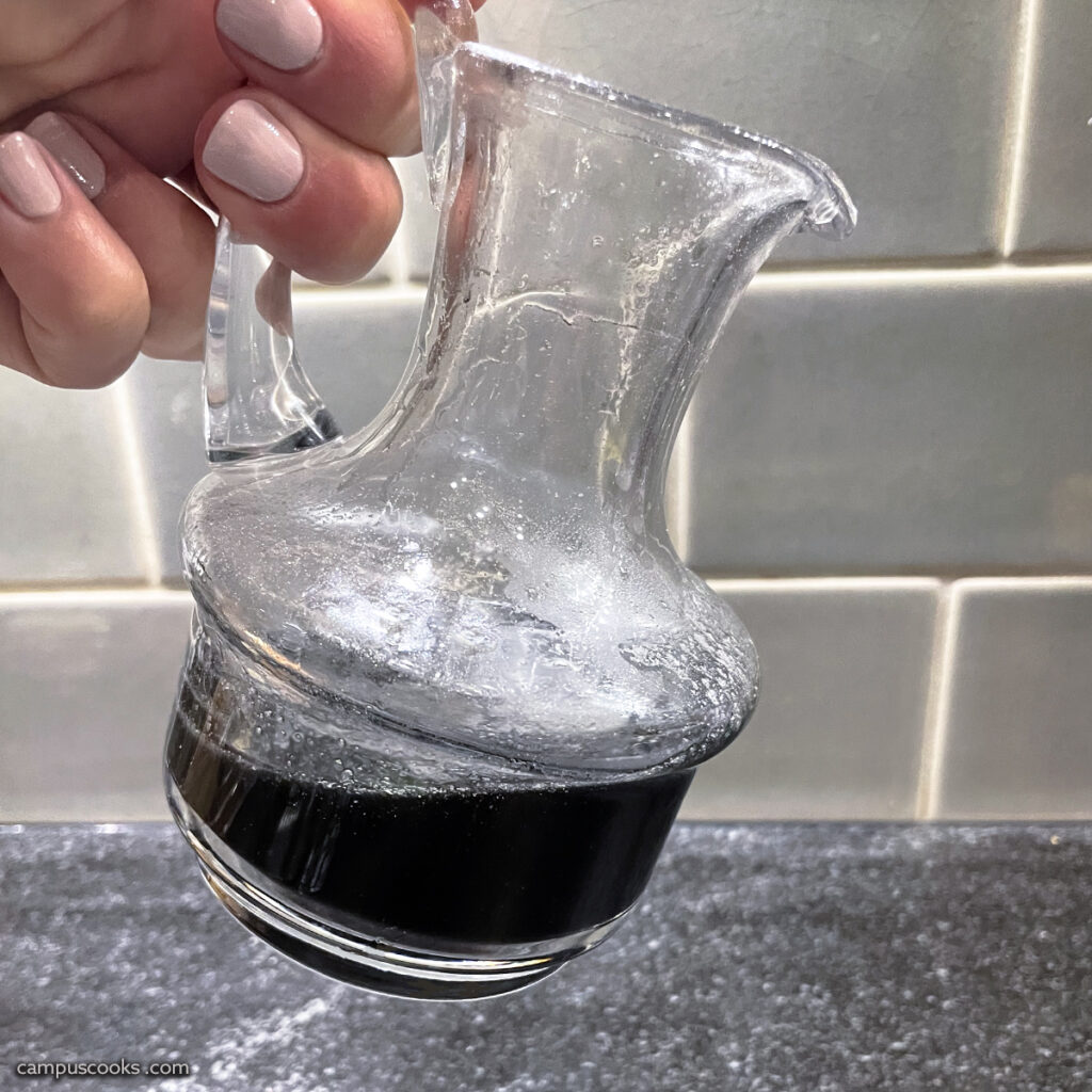 a well-manicured hand holds a small glass pitcher, filled with a black syrup over a black granite countertop.